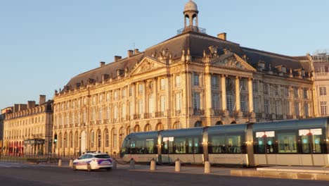 Place-De-La-Bourse-En-Burdeos-Al-Amanecer-Con-El-Paso-Del-Tranvía-Y-Algunos-Autos,-Con-El-Museo-Francés-De-Aduanas-En-La-Parte-De-Atrás