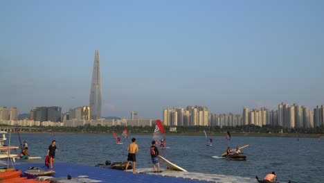 Gente-Coreana-Caminando-En-El-Muelle-Flotante-Mientras-Otros-Practican-Windsurf-En-El-Río-Han-Junto-A-La-Torre-Lotte-World,-Seúl-Al-Atardecer