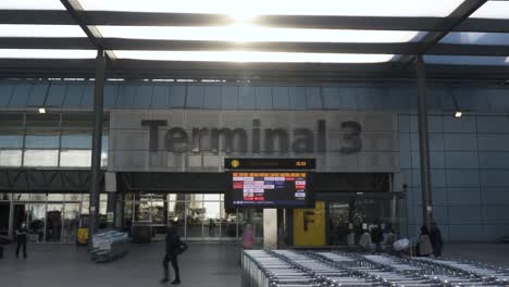 Fila-De-Carros-De-Equipaje-Fuera-Del-Edificio-De-La-Terminal-3-De-Heathrow-Con-Tablero-De-Salida-De-La-Aerolínea-Con-El-Sol-Brillando-A-Través-Del-Voladizo-Del-Techo