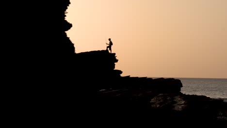 Mann-Mit-Angelrute-über-Großen-Felsigen-Klippen-Am-Strand-Bei-Sonnenuntergang,-Mittlere-Handaufnahme