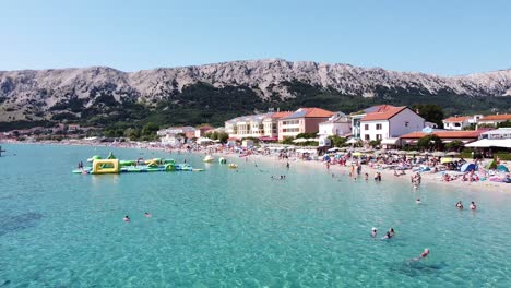 Playa-Baska,-Isla-Krk,-Croacia---Vista-Aérea-De-Drones-Del-Bulevar,-Turistas,-Tumbonas-Y-Juegos-Acuáticos-Durante-Las-Vacaciones-De-Verano