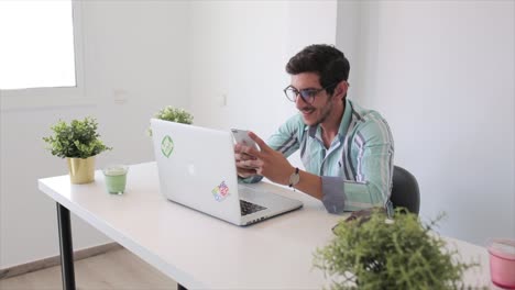 A-static-shot-of-a-happy-man-in-glasses-with-laptop-computer-working-at-office-and-surfing-his-mobile