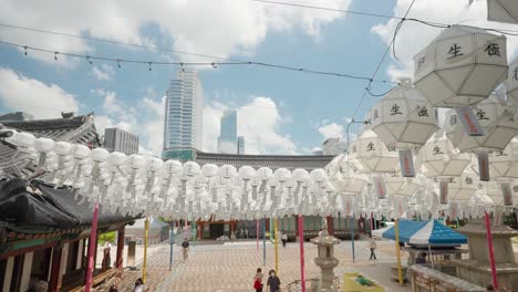 Personas-Con-Mascarillas-Protectoras-Que-Visitan-El-Templo-Bongeunsa-Decorado-Con-Farolillos-Blancos-De-Papel-Redondos-Clásicos-Colgantes-Con-Tarjetas-De-Bendición-En-Un-Día-Soleado-Con-Nubes