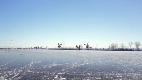 Gente-Patinando-Sobre-Hielo-En-Un-Canal-Holandés-Congelado,-Molinos-De-Viento-En-El-Fondo,-Escena-De-Invierno