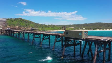 Male-jumping-off-pier-into-sea-with-friends-watching,-aerial-reveal