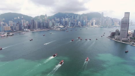 Convoy-of-local-Fishing-boats-causing-in-Hong-Kong-Victoria-bay,-with-city-skyline-in-the-horizon,-Aerial-view