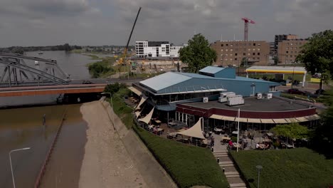 Luftaufnahmen,-Die-Sich-Seitwärts-Drehen-Und-Das-Restaurant-Und-Firmengebäude-Ijsselpaviljoen-Am-Fluss-Ijssel-Mit-Menschen-Auf-Der-Terrasse-Bei-Hohem-Wasserstand-Zeigen