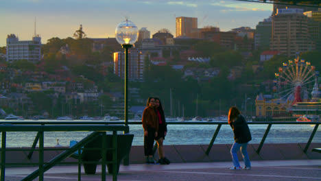 Amigas-Tomando-Fotos-Juntas-En-El-Paseo-Marítimo-Del-Puerto-De-Sydney-En-Nsw,-Australia-Al-Atardecer