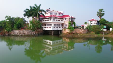 Aerial-slow-motion-drone-movement-of-a-three-story-house-with-luxurious-balconies-and-palm-trees-reflected-in-the-blue-water-lake,-in-Vadodara,-India