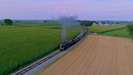 Una-Vista-Aérea-De-Un-Tren-De-Vapor-Que-Se-Acerca-Volando-Viajando-A-Través-De-Tierras-De-Cultivo-Y-Campos-De-Maíz-Que-Soplan-Humo-En-Un-Día-Soleado-De-Verano