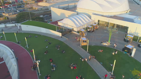 Video-Aéreo-De-Drones-De-La-Terraza-Al-Aire-Libre,-Personas-Relajadas-Sentadas-En-Césped-Sintético-Verde,-Escalofriante,-Fuera-Del-Centro-Comercial-De-Costa-Rica,-Video-De-Drones-En-El-Centro-Comercial-Al-Atardecer,-Al-Aire-Libre,-Concierto-Al-Aire-Libre