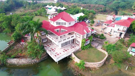 Vista-Aérea-Giratoria-De-Drones-De-Una-Lujosa-Casa-De-Vacaciones,-Para-Relajarse-Y-Disfrutar-Nadando-En-El-Lago-Y-La-Piscina,-Junto-A-Las-Palmeras-Y-La-Vegetación-Original-De-Vadodara,-India