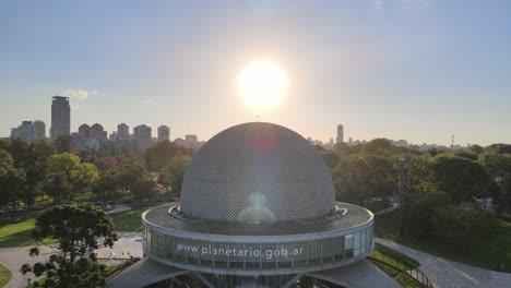 Pedestal-Aéreo-Arriba,-Con-Vista-Al-Hermoso-Sol-Brillante-Que-Brilla-Detrás-De-La-Famosa-Estructura-Del-Edificio-Planetario-Galileo-Galilei-En-Los-Bosques-De-Palermo,-Centro-De-Buenos-Aires