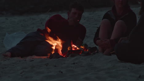 Group-of-Young-People-Warming-Around-A-Bonfire-at-Dusk-on-Beach,-Cornwall,-England,-UK---telephoto