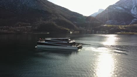 Electric-passenger-boat-Vision-of-the-fjords-sailing-backwards-after-showing-tourists-a-local-waterfall-in-Flam---Static-aerial-with-sunlight-reflections-in-sea-surface-and-on-boat