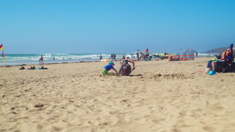 Amorosa-Madre-E-Hijo-Jugando-Juntos-Cavando-Arena-De-Playa-En-Perranporth,-Cornualles,-Inglaterra---Plano-General