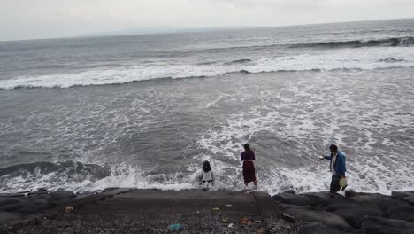 Mujer-Balinesa-Rezando-En-El-Mar-Después-De-Una-Ceremonia-Ritual-Hindú-De-Melukat,-Playa-De-Bali-De-Arena-Negra-Volcánica-Que-Representa-Una-Oración-Para-Adorar-A-Los-Dioses,-Masceti,-Gianyar