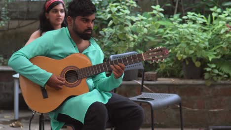 Hombre-Adulto-Joven-Tocando-Una-Canción-En-La-Guitarra,-Camisa-Turquesa-Brillante,-Chica-Con-Un-Pañuelo-En-El-Fondo