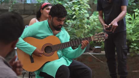 Joven-Adulto-Tocando-Una-Canción-En-La-Guitarra,-Camisa-Turquesa-Brillante,-Chica-Con-Un-Pañuelo-En-El-Fondo-Y-Un-Baterista-En-Primer-Plano