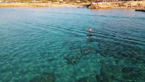 Las-Imágenes-De-Un-Dron-Siguen-A-Una-Persona-Que-Navega-En-Una-Tabla-Flotante-Sobre-Las-Aguas-Cristalinas-Y-Azules-De-Un-Océano-Cerca-De-La-Costa