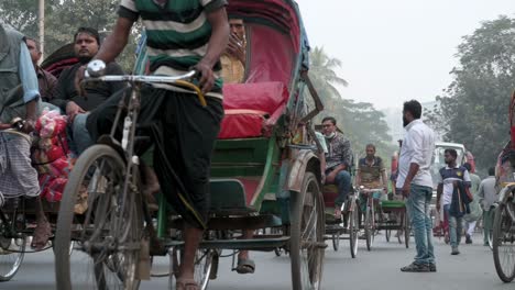 Zeitlupenaufnahme-Von-Pendlern-Aus-Bangladesch,-Die-Auf-Rikschas-Und-Fahrzeugen-Fahren,-Während-Fußgänger-Versuchen,-Eine-Stark-Befahrene-Straße-In-Dhaka-Zu-überqueren