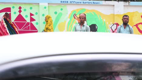 Bangladeshi-rickshaw-pullers-stand-on-the-sidewalk-of-a-busy-and-colorful-road-as-vehicles-drive-past-them-in-Dhaka,-Bangladesh