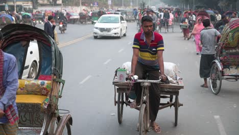 Zeitlupenaufnahmen-Von-Bangladeschischen-Pendlern,-Die-Mit-Anderen-Fahrzeugen-Auf-Rikschas-Auf-Einer-Stark-Befahrenen-Straße-Fahren,-Während-Im-Hintergrund-Ein-Fußgänger-Die-Straße-überquert