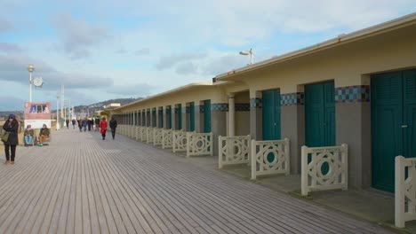 Menschen,-Die-Auf-Einer-Holzallee-Neben-Den-Berühmten-Normannischen-Strandhütten-Und-Der-Holzpromenade-Les-Planches-Spazieren,-Die-Mit-Namen-Amerikanischer-Filmschauspieler-Und-Regisseure-In-Deauville,-Frankreich,-Bemalt-Ist