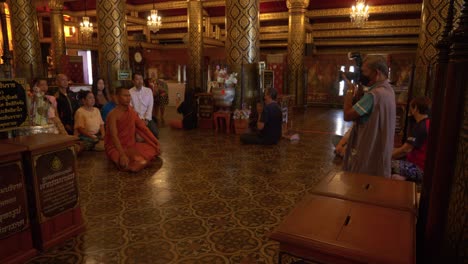 Photographer-taking-pictures-of-monk-and-tourists-praying-to-Buddha-inside-Buddhist-temple,-Thailand