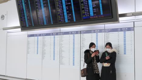 Reisepassagiere-Nutzen-Ihre-Smartphones-Neben-Einem-Bildschirm,-Der-Anzeigt,-Wo-Sich-Die-Check-in-Schalter-Der-Fluggesellschaften-Am-Internationalen-Flughafen-Chek-Lap-Kok-In-Hongkong-Befinden