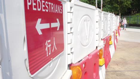 Vista-Lateral-De-La-Señalización-Roja-De-Peatones-En-La-Barrera-En-El-Sitio-De-Obras-De-Construcción-En-Tseung-Kwan-O,-Hong-Kong-Con-Gente-Caminando-En-Segundo-Plano