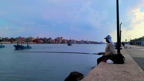 Barcos-Que-Pasan-En-Un-Día-Nublado-Mientras-Un-Pescador-Egipcio-Pesca-Con-Una-Caña-De-Pescar-Larga-En-Ras-El-bar,-Donde-El-Río-Nilo-Se-Encuentra-Con-El-Mar-Mediterráneo