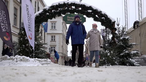 In-slow-motion-a-couple-walk-under-an-archway-made-of-pine-branches-at-the-entrance-to-a-Christmas-fair-of-festive-stalls-in-a-snow-covered-town-square