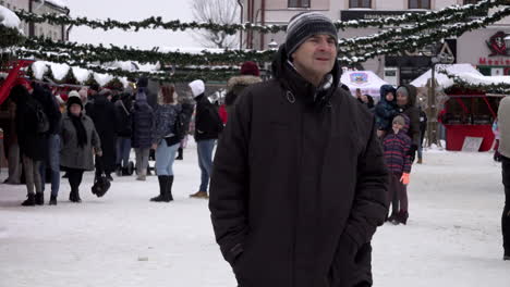 Ein-Mann-Fotografiert-Seine-Familie-Auf-Seinem-Smartphone,-Während-Menschen-Einen-Weihnachtsmarkt-Mit-Festlichen-Ständen-Auf-Einem-Schneebedeckten-Stadtplatz-Besuchen