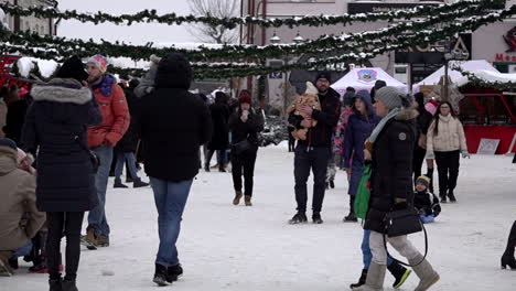 In-Zeitlupe-Besuchen-Männer,-Frauen-Und-Kinder-Einen-Weihnachtsmarkt-Mit-Festlichen-Ständen-Auf-Einem-Schneebedeckten-Stadtplatz