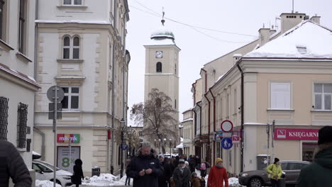 Männer,-Frauen-Und-Kinder-In-Winterkleidung-Laufen-Durch-Schneebedeckte-Straßen,-Umgeben-Von-Idyllischen-Gebäuden-Und-Einem-Glockenturm