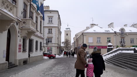 Hombres,-Mujeres-Y-Niños-Vestidos-De-Invierno-Caminan-Por-Calles-Cubiertas-De-Nieve-Rodeadas-De-Edificios-Idílicos-Y-Una-Torre-De-Reloj