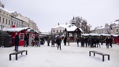 Männer,-Frauen-Und-Kinder-Besuchen-Einen-Weihnachtsmarkt-Mit-Festlichen-Ständen-Auf-Einem-Schneebedeckten-Stadtplatz,-Umgeben-Von-Idyllischen-Gebäuden