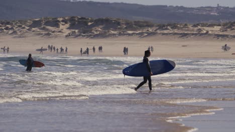 Surfer-Geht-Im-Seichten-Meerwasser-Vom-Strand-Weg,-Menschen-Im-Hintergrund