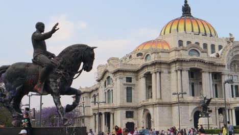 Una-Toma-Completa-Con-Seguimiento-De-Un-Monumento-De-Un-Hombre-A-Caballo-Con-El-Palacio-De-Bellas-Artes-En-La-Ciudad-De-México,-Con-Algunas-Personas-Caminando-En-Una-Tarde-Clara