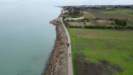 Family-riding-bicycles-at-ocean-shore-near-a-town-farm,-Aerial-dolly-out-shot