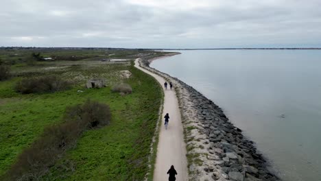 Four-people-riding-bicycles-around-the-road-near-the-ocean,-Aerial-follow-shot