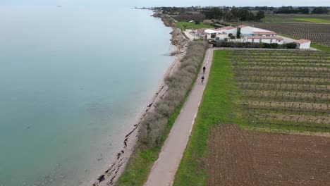 Two-women-ride-bicycles-near-farmland-and-stop-to-see-the-coastline,-Aerial-orbit-around-shot