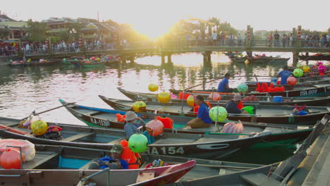 Vista-Cinematográfica-Del-Puente-Peatonal-Con-Gente-Caminando-Sobre-El-Río-Con-Botes-Tradicionales-Con-Jinetes-Flotando-En-El-Río-Al-Atardecer-En-El-Casco-Antiguo-De-Hoi-An,-Vietnam