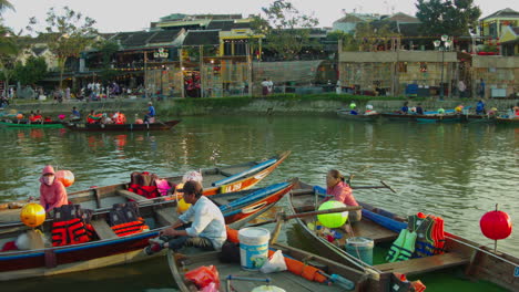 Jinetes-De-Botes-Sampán-Esperando-Clientes-En-Sus-Botes-En-El-Río-Thu-Bon-De-La-Antigua-Ciudad-De-Hoi-An-Con-Casas-Tradicionales-De-Bambú-Como-Telón-De-Fondo,-Vietnam