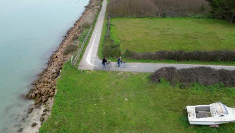 Menschen,-Die-Mit-Dem-Fahrrad-An-Einer-Kurve-In-Der-Nähe-Eines-Bootes-Und-Des-Ozeans-Im-Dorf-Fahren,-Aufnahme-Aus-Der-Luft