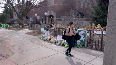 Edificio-Sindical-En-El-Campus-De-La-Universidad-Estatal-De-Michigan,-Lugar-De-Un-Tiroteo-Masivo-El-13-De-Febrero-De-2023-Con-Un-Monumento-Floral-Y-Una-Joven-Caminando