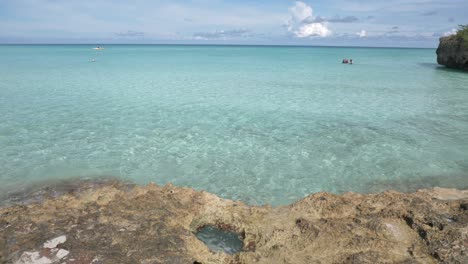 Orilla-Rocosa-Golpeada-Por-Las-Olas-Y-La-Gente-Bañándose-En-El-Océano-Azul