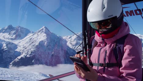 Mujer-En-Traje-De-Esquí-Enviando-Mensajes-De-Texto-En-Un-Teléfono-Inteligente-Dentro-De-Un-Teleférico-De-Una-Estación-De-Esquí-De-Montaña-En-Un-Día-Soleado