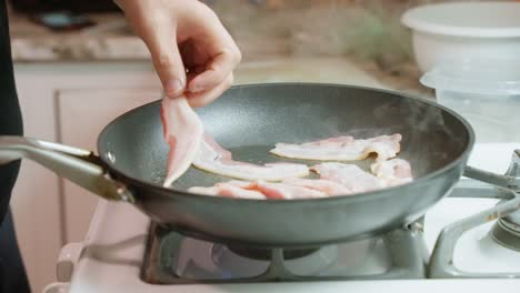 Person-cooking-bacon-and-adding-additional-half-slices-in-a-Teflon-frying-pan-with-a-spatula-on-a-gas-stove